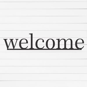 May include: Black metal "welcome" sign in a classic font, mounted on a white, horizontal-slat wooden background. The sign is a single line, with the letters connected at the base.