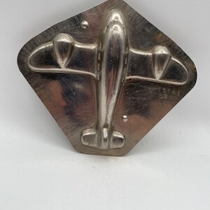 Op de afbeelding: Een zilveren metalen mal in de vorm van een vliegtuig. De mal heeft een gedetailleerd ontwerp van een propeller vliegtuig met vleugels, een romp en een staart.