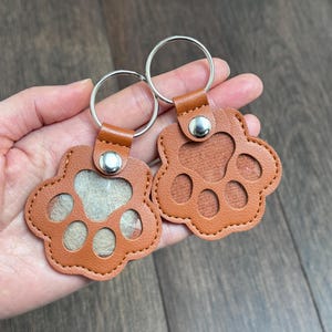 May include: Two brown leather paw-shaped keychains with silver key rings. The keychains have a stitched border and a snap closure. One keychain has a clear window, and the other has a solid brown background.