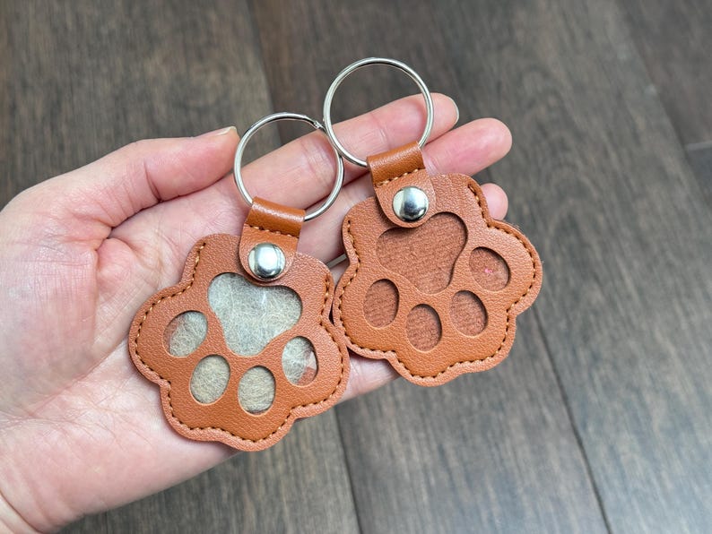 May include: Two brown leather paw-shaped keychains with silver key rings. The keychains have a stitched border and a snap closure. One keychain has a clear window, and the other has a solid brown background.