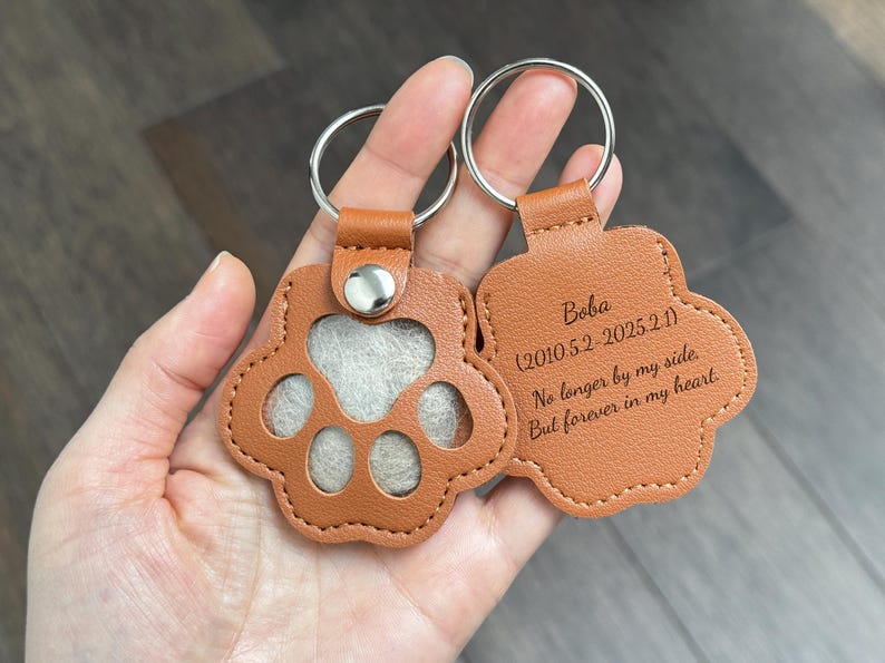 May include: A tan leather paw-shaped keychain with a silver keyring. The front has a paw print cutout with a light gray insert. The back has engraved text: "Boba (2010.5.2-2025.2.1) No longer by my side. But forever in my heart."