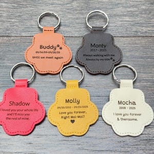 May include: Five paw-shaped keychains in various colors: peach, gray, red, yellow, and cream. Each keychain has a silver ring and personalized text, including names and dates. The keychains are arranged on a wooden surface.
