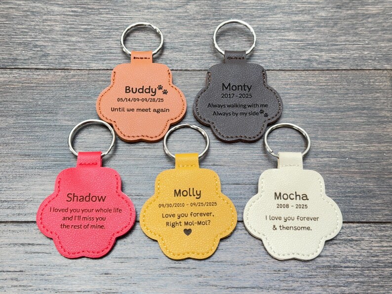 May include: Five paw-shaped keychains in various colors: peach, gray, red, yellow, and cream. Each keychain has a silver ring and personalized text, including names and dates. The keychains are arranged on a wooden surface.
