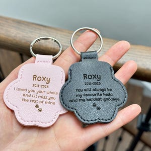 May include: Two paw-shaped keychains, one pink and one gray, held in a hand. Each keychain has a silver ring and is engraved with the name "Roxy" and dates, along with a heartfelt message and paw prints.