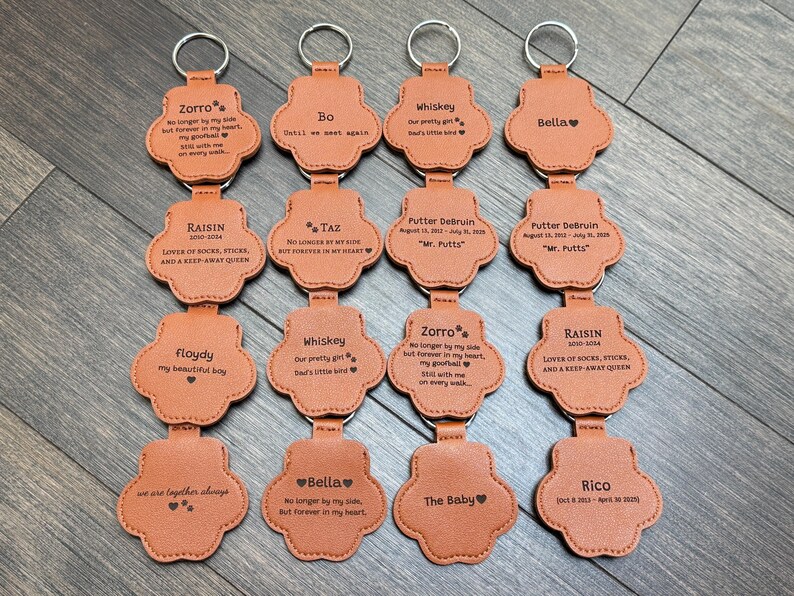 May include: A collection of personalized paw-shaped keychains in a light brown color. Each keychain has a silver keyring and features engraved text with names and dates. The keychains are arranged on a wooden surface.