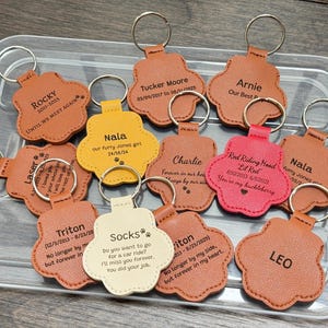 May include: A collection of personalized paw-shaped keychains in various colors, including brown, yellow, and red. Each keychain features a name and dates, with phrases like "Forever in our hearts" and "Until we meet again."