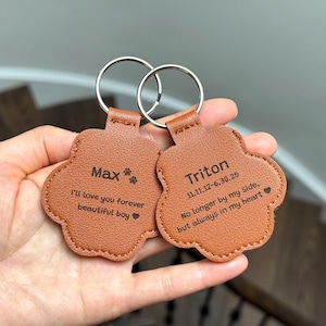 May include: Two brown leather paw-shaped keychains with silver key rings. One keychain reads "Max" with "I'll love you forever beautiful boy" below. The other reads "Triton" with dates and a sentimental message.