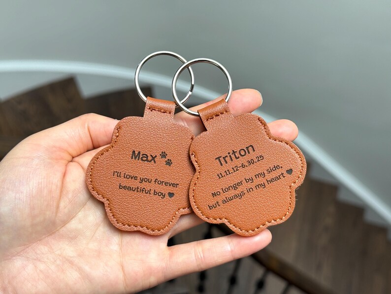 May include: Two brown leather paw-shaped keychains with silver key rings. One keychain reads "Max" with "I'll love you forever beautiful boy" below. The other reads "Triton" with dates and a sentimental message.