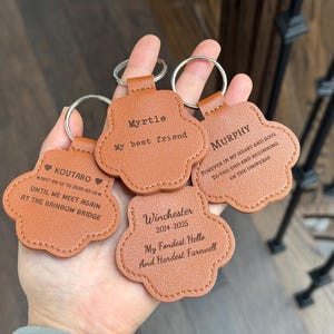May include: Four personalized paw-shaped keychains in a light brown color. Each keychain has a silver ring and features engraved text with names and dates. The keychains are a memorial item.