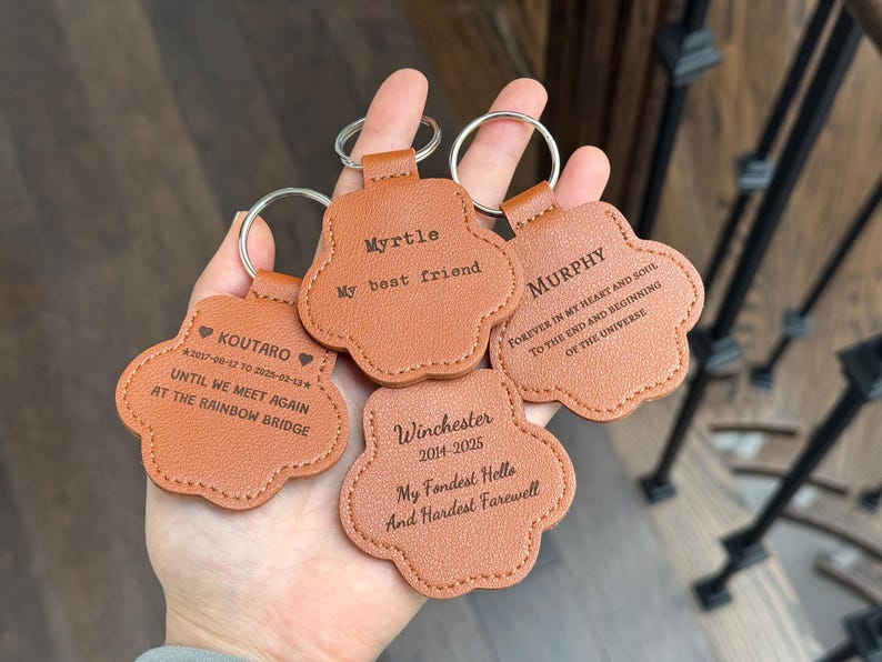 May include: Four personalized paw-shaped keychains in a light brown color. Each keychain has a silver ring and features engraved text with names and dates. The keychains are a memorial item.
