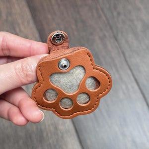 May include: A brown leather paw print-shaped keychain with a gray felt center and a snap closure. The keychain is designed with stitched edges and a small loop for attachment.