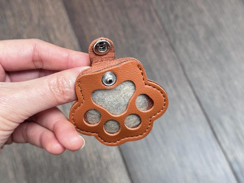 May include: A brown leather paw print-shaped keychain with a gray felt center and a snap closure. The keychain is designed with stitched edges and a small loop for attachment.