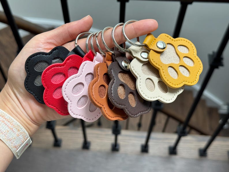 May include: A collection of colorful paw-shaped keychains in black, red, pink, brown, and yellow. Each keychain has a silver ring and a leather strap. The paw prints are stitched with a contrasting thread.