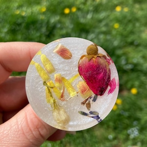 May include: A round, clear resin paperweight with pressed flowers and petals inside. The flowers are pink, yellow, and purple.