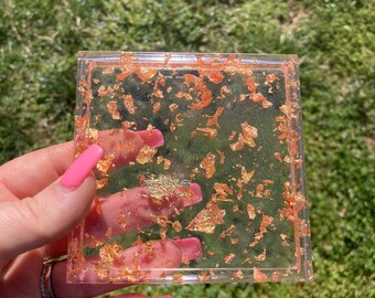 May include: A square clear resin tray with gold flakes embedded inside. The tray is translucent and has a light pink hue.