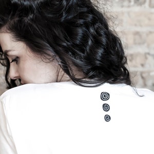 May include: A white t-shirt featuring three black spiral motifs on the back. The model has dark, curly hair. The background is a light-coloured brick wall.