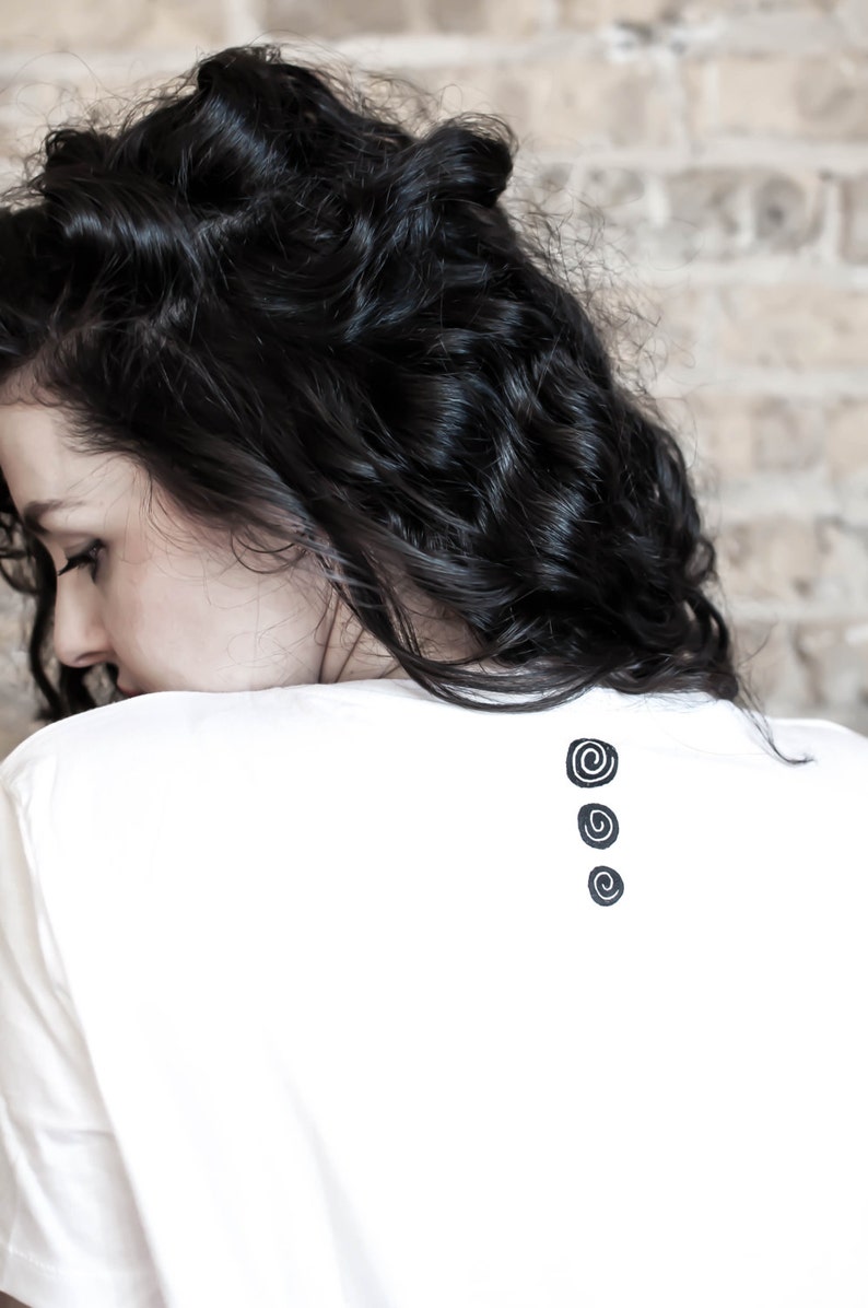 May include: A white t-shirt featuring three black spiral motifs on the back. The model has dark, curly hair. The background is a light-coloured brick wall.
