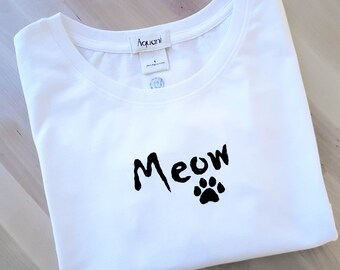 May include: White t-shirt with the word "Meow" in a handwritten style and a black paw print. The shirt has a crew neck and a tag that reads "Aquani". The shirt is folded on a light wood surface.