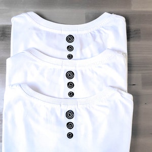 May include: Three folded white t-shirts with a crew neck and a black spiral design down the centre. The shirts are stacked, with the top shirt slightly offset. The background is a light wood grain pattern.