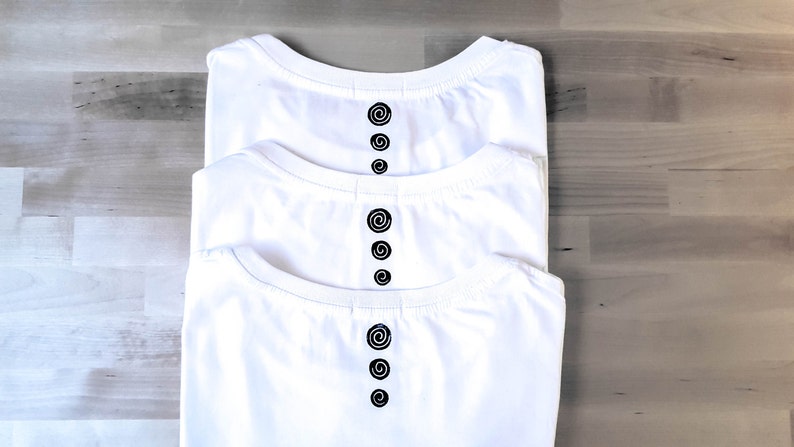 May include: Three folded white t-shirts with a crew neck and a black spiral design down the centre. The shirts are stacked, with the top shirt slightly offset. The background is a light wood grain pattern.