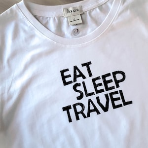 May include: White crew neck t-shirt with the words "EAT SLEEP TRAVEL" printed in black. The shirt has a tag at the neckline and a small button detail. The fabric appears to be soft and lightweight.