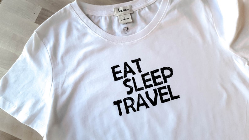May include: White crew neck t-shirt with the words "EAT SLEEP TRAVEL" printed in black. The shirt has a tag at the neckline and a small button detail. The fabric appears to be soft and lightweight.