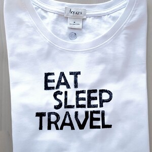 May include: White t-shirt with the words "EAT SLEEP TRAVEL" printed in bold black letters. The shirt has a crew neck and a small tag at the neckline. The shirt is folded neatly.