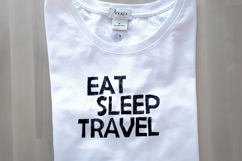 May include: White t-shirt with the words "EAT SLEEP TRAVEL" printed in bold black letters. The shirt has a crew neck and a small tag at the neckline. The shirt is folded neatly.