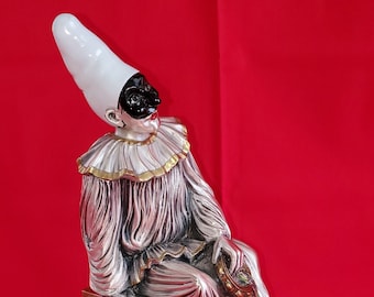 Op de afbeelding: Een zilveren beeldje van een nar met een witte hoed en een zwart masker, zittend op een houten kruk met een hartvormige uitsparing. De nar houdt een klein, versierd voorwerp in zijn hand. Het beeldje staat op een zwarte basis.