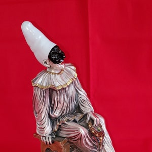 May include: A silver figurine of a jester wearing a white hat and a black mask, sitting on a wooden stool with a heart-shaped cutout. The jester is holding a small, ornate object in their hand. The figurine is on a black base.