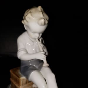 May include: A porcelain figurine of a young boy playing a flute, sitting on a wooden box with two small birds at his feet. The figurine is white with a light brown glaze on the boy's hair and the wooden box.