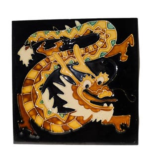 May include: Square ceramic tile featuring a detailed dragon design. The dragon is depicted in shades of brown, yellow, and white against a black background. The tile has a glossy finish and is likely intended for decorative use.