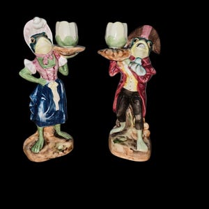 May include: Two ceramic frog figurines, one in a pink and blue dress and the other in a red coat, each holding a white candle holder. The figurines are on a brown base and have a whimsical, antique aesthetic.