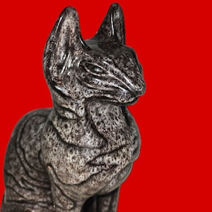 Puede incluir: Figura de gato de cerámica gris oscuro con acabado moteado. El gato está sentado con las orejas erguidas, sobre un fondo rojo vibrante. La figura tiene una superficie texturizada, lo que añade profundidad a su diseño.