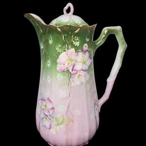 May include: A vintage porcelain coffee pot with a lid, featuring a green and pink floral design. The pot has a gold trim and a branch-like handle. The floral pattern includes pink and purple flowers with green leaves.