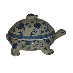 Pode incluir: Uma caixa de cerâmica em forma de tartaruga com tampa. A caixa é decorada com um padrão floral azul e verde. A tampa tem uma tartaruga menor no topo.