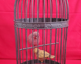 May include: A black metal birdcage with a decorative design around the base. Two small ceramic birds are inside the cage, one yellow and one red.