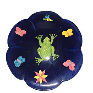 Può includere: Un piatto in ceramica blu scuro a forma di fiore con una rana dipinta, farfalle, un'ape e un motivo a stella. La rana è verde con macchie gialle e le farfalle sono rosa, blu e gialle.