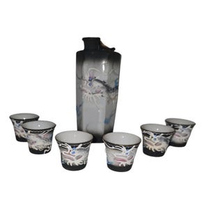 May include: A ceramic sake set consisting of one tall, hexagonal bottle and six small cups. The pieces feature a white base with a black gradient at the rims and bases, decorated with raised, detailed white dragon motifs and blue accents.