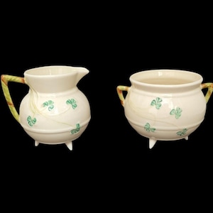 Puede incluir: Juego de jarra de crema y azucarero de cerámica color crema. La jarra de crema tiene un pico y un asa estampada. El azucarero tiene dos asas. Ambas piezas están decoradas con diseños de tréboles verdes y se asientan sobre tres patas pequeñas.