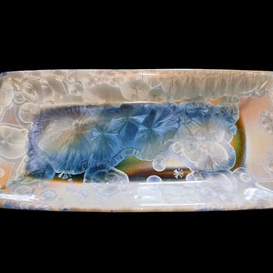 May include: Rectangular ceramic serving dish with a unique crystalline glaze. The dish features a central area of blue and green hues, surrounded by white and tan tones. The glaze has a textured, almost bubbly appearance, with a small star-like design.