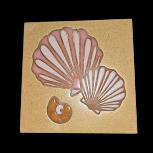 May include: A square ceramic tile with a speckled beige background featuring a large pink scallop shell, a smaller white scallop shell, and a tiny orange spiral seashell. The shells are outlined in dark brown, creating a distinct graphic design.