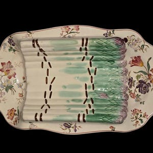 May include: A ceramic asparagus serving dish with a scalloped edge, featuring a central molded relief of green and white asparagus stalks tied with brown bands, surrounded by a cream-colored border decorated with colorful floral patterns.