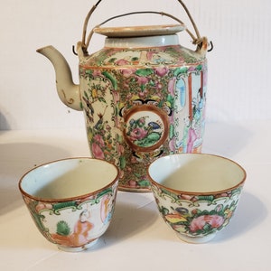 Könnte beinhalten: Ein antikes chinesisches Teeset mit einer Teekanne und zwei Tassen. Die Teekanne hat einen Drahtgriff und ein florales Design in Rosa, Grün und Blau. Die Tassen haben ein ähnliches florales Design.