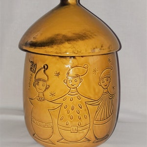 Könnte beinhalten: Eine Keramik-Keksdose mit einem Deckel in Form eines Pilzes. Die Dose ist mit drei Comicfiguren in Braun und Weiß verziert und hat eine braune Glasur.