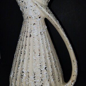 May include: A white ceramic pitcher with a textured surface and gold accents. The pitcher has a wide mouth and a curved handle.
