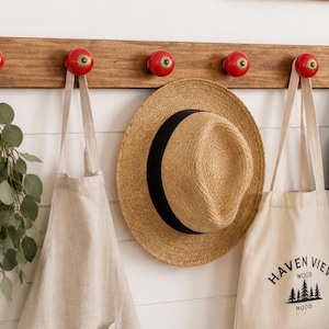 May include: A horizontal wooden wall rack with five red ceramic knobs. Hanging from the knobs are a bunch of green eucalyptus, a beige apron, a straw hat with a black band, a beige canvas tote bag with the text Haven View Wood Mood, and a denim jacket.