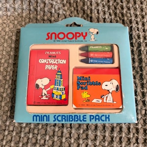 May include: A vintage Snoopy Mini Scribble Pack featuring a light blue box with Snoopy and Woodstock graphics. The pack includes mini construction paper, a scribble pad, and three crayons. The packaging reads "Mini Scribble Pack."