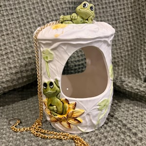 May include: White ceramic frog-themed bird feeder with a gold chain. The feeder features two green frogs, one resting on top and another perched on a yellow flower. The feeder has a large opening and is decorated with leaf and flower details.