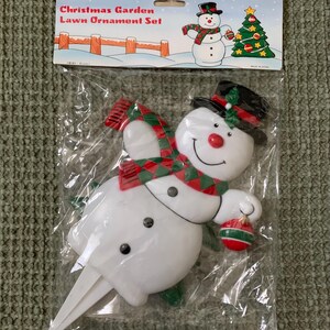 May include: A Christmas Garden Lawn Ornament Set in a clear plastic package. The set includes a white snowman with a black top hat, red scarf, and a red nose. The packaging has the words "Christmas Garden Lawn Ornament Set".